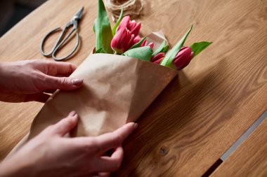 Ahşap masa üzerinde pembe lale buketi düzenleyen kadın elleri, çiçeklerle dolu iş yeri, iş yeri, diy, bahar hediyesi konsepti yukarıdan.