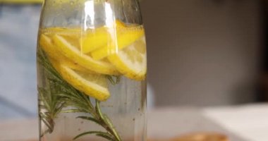 Kadın eli cam şişeye limon koyuyor, kadın hazırlanıyor, limon ve biberiyeyle sağlıklı sudan arınıyor, evde beyaz bir masada taze limonata içiyor, yaz içkisi içiyor.