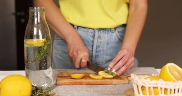 女性の手でレモンをカット 女性の準備 レモンとローズマリーとデトックス健康的な水を作る 自宅で白いテーブルの上にガラス瓶で新鮮なレモネード 夏の飲み物 — ストック動画
