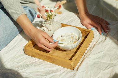 Kot pantolonlu bir kadın yatakta oturuyor ve sabah güneşinde sağlıklı tahıl gevreği kasesi yiyor..