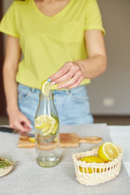 Kadın eli cam şişeye limon koyuyor, kadın hazırlanıyor, limon ve biberiyeyle sağlıklı sudan arınıyor, evde beyaz bir masada taze limonata içiyor, yaz içkisi içiyor.