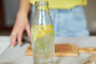 Kadın eli cam şişeye limon koyuyor, kadın hazırlanıyor, limon ve biberiyeyle sağlıklı sudan arınıyor, evde beyaz bir masada taze limonata içiyor, yaz içkisi içiyor.