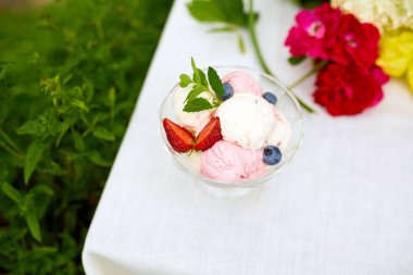 Lezzetli dondurma ve taze yaban mersini, kasede çilek, yaz mevsiminde bir bahçede bir masada çiçekle sunuluyor.