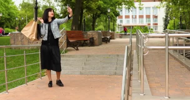 Morena mulher segurando sacos de compras de papel e tirando selfie por smartphone no parque — Vídeo de Stock