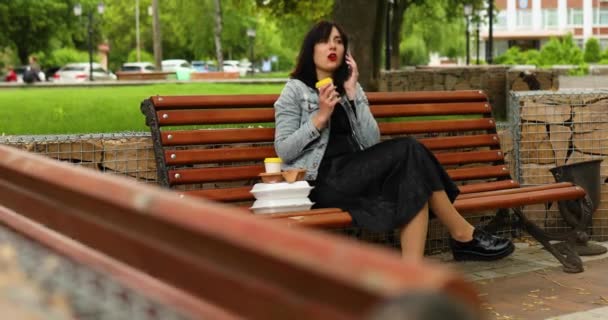 女人坐在公园的长椅上 一边用手机聊天 一边带走食物和咖啡 一边吃午饭 — 图库视频影像