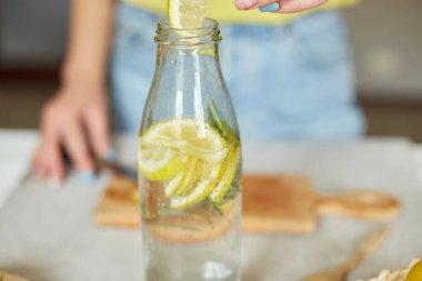 Kadın eli cam şişeye limon koyuyor, kadın hazırlanıyor, limon ve biberiyeyle sağlıklı sudan arınıyor, evde beyaz bir masada taze limonata içiyor, yaz içkisi içiyor.