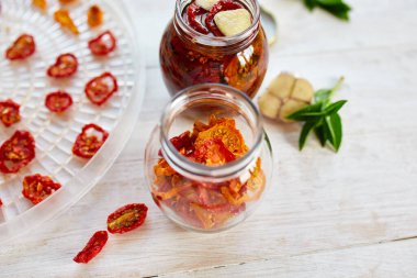 Ev yapımı güneşte kurutulmuş domatesler, beyaz ahşap zemin üzerinde cam bir kavanozda zeytinyağında sarımsak. Uzayı kopyala, kapat. Mutfak için yazdır.