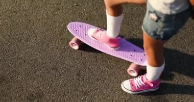 Kaykay kullanmayı öğrenen küçük bir kız, beyaz çoraplar ve pembe ayakkabılar giyen isimsiz bir çocuk yaz şehrinin kırsal kesiminde kaykaya biniyor..