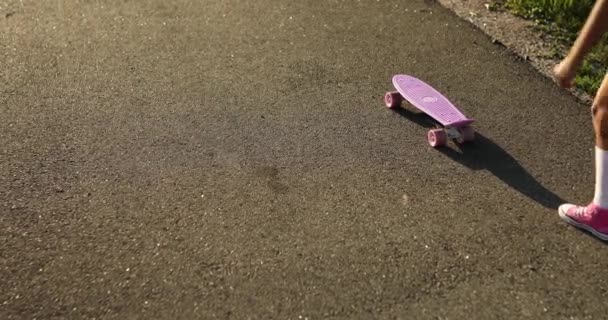 スケートボードを使うことを学ぶ小さな女の子 白い靴下とピンクの靴を着た匿名の子供夏の街の田舎道でスケートボードに乗る — ストック動画