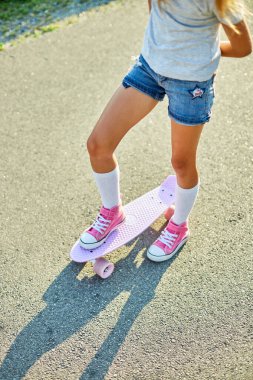 Kaykay kullanmayı öğrenen küçük bir kız, beyaz çoraplar ve pembe ayakkabılar giyen isimsiz bir çocuk yaz şehrinin kırsal kesiminde kaykaya biniyor..