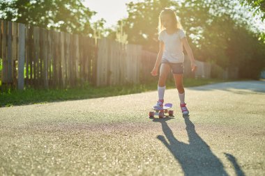 Kaykay kullanmayı öğrenen küçük bir kız, beyaz çoraplar ve pembe ayakkabılar giyen isimsiz bir çocuk yaz şehrinin kırsal kesiminde kaykaya biniyor..