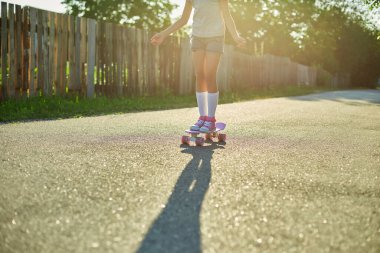 Kaykay kullanmayı öğrenen küçük bir kız, beyaz çoraplar ve pembe ayakkabılar giyen isimsiz bir çocuk yaz şehrinin kırsal kesiminde kaykaya biniyor..