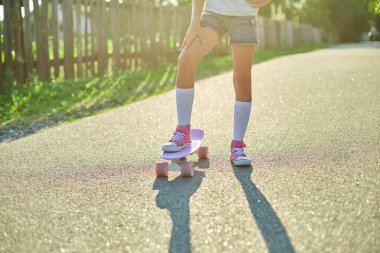 Kaykay kullanmayı öğrenen küçük bir kız, beyaz çoraplar ve pembe ayakkabılar giyen isimsiz bir çocuk yaz şehrinin kırsal kesiminde kaykaya biniyor..