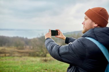 Sırt çantalı gezgin, akıllı telefonuyla fotoğraf ya da selfie çeker. Telefonu nehir ve dağlarla bulanık arkaplan, yaşam tarzı..