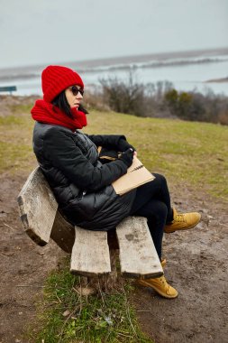 Siyah ceketli, kırmızı şapkalı gezgin kadın deftere bir şeyler yazıyor, not alıyor, nehir manzarasına karşı eskizler çiziyor, yerel seyahat, akıl sağlığı. Sonbahar, sonbahar