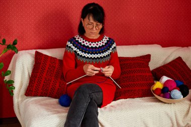 Olgun bir kadın rahat bir kanepede oturup örgü örerken hafta sonunun tadını çıkarıyor, kıdemli kadın örgü örüyor, evde dinleniyor hafta sonları en sevdiği hobi aktivitelerini yapıyor, refah kavramını geliştiriyor.