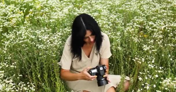 Kadın fotoğrafçı çiçek tarlasında oturuyor ve kameraya bakıyor. — Stok video