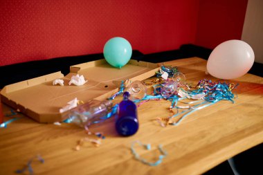 Parti kutlamalarından sonra parti kaosundan sonra parti artıklarını kutlamak için pizza kutusu, boş mavi şişeler, şarap bardağı ve parti süslemeleri olan dağınık ahşap masa.