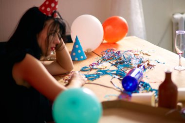 Kırmızı rujlu ve şapkalı genç bir kadın doğum günü partisinden sonra dağınık bir odada yorgun bir şekilde oturuyor.