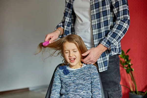 Father combing, brushing his daughter\'s hair at home, child making faces about hair pulling, angry girl feeling pain, family moments, spending time together.