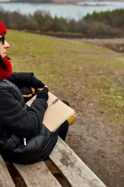 Siyah ceketli, kırmızı şapkalı gezgin kadın deftere bir şeyler yazıyor, not alıyor, nehir manzarasına karşı eskizler çiziyor, yerel seyahat, akıl sağlığı. Sonbahar, sonbahar