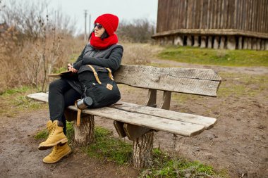 Siyah ceketli, kırmızı şapkalı gezgin kadın deftere bir şeyler yazıyor, not alıyor, nehir manzarasına karşı eskizler çiziyor, yerel seyahat, akıl sağlığı. Sonbahar, sonbahar