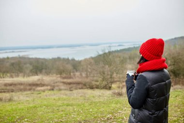 Kırmızı şapkalı ve eşarplı kadın, metal bardaktan çay içiyor, seyahat konsepti, sonbahar manzarası.