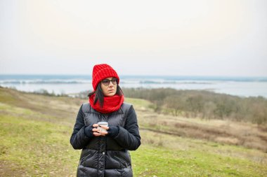 Kırmızı şapkalı ve eşarplı kadın, metal bardaktan çay içiyor, seyahat konsepti, sonbahar manzarası.