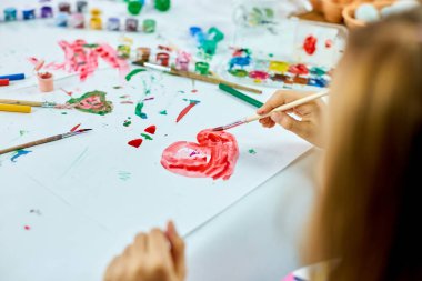 Masada oturan tatlı çocuk beyaz kağıda kırmızı kalp çiziyor, sanat okulu konsepti, evde suluboya resimlerle resim yapan kız çocuğu.