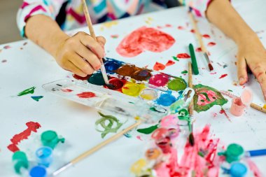 Masada oturan tatlı çocuk beyaz kağıda kırmızı kalp çiziyor, sanat okulu konsepti, evde suluboya resimlerle resim yapan kız çocuğu.