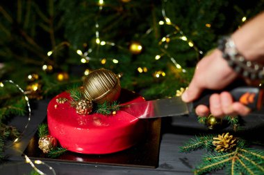 Bir el, neşeli bir tatil atmosferinde yanıp sönen ışıklarla çevrili altın süsler ve çam ağacıyla süslenmiş canlı kırmızı pastayı dilimlemek için bir bıçak tutuyor..