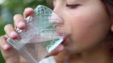 Bir kız bahçede duruyor ve bardaktan su içiyor. O, güneşli günün ve etrafındaki yeşilliğin tadını çıkarıyor. Suyun serinliği onu canlandırıyor..