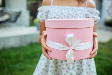 Bahçede elinde kurdeleli pembe bir hediye kutusu olan genç bir kız duruyor. Çiçekli bir elbise giyiyor ve çevresi yeşilliklerle çevrili ve öğleden sonra boyunca arka planda bir evi var..