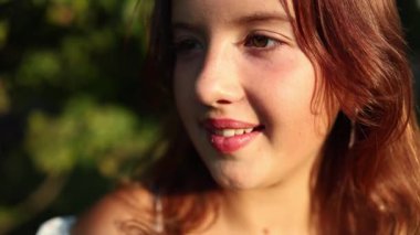 Yakından bakınca genç bir kız gülümsüyor açık havada güneşli bir günün tadını çıkarırken, öğleden sonra aydınlığında yeşilliklerle çevrili.