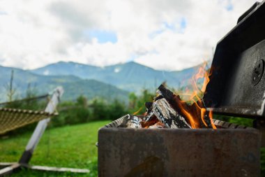 Izgarada bir kamp ateşi yanıyor ve odunlar alev alıyor. Arka planda, açık bir gökyüzünün altında yeşil dağlar var. Huzurlu bir yerde sıcak bir gün.