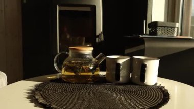 Çay, içinde bitkiler olan cam bir çaydanlıkta demlenir. İki beyaz kupa çaydanlığın yanındaki dairesel bir paspasın üzerinde dinleniyor. Sabah odaya ışık yağar..