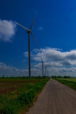 Mavi gökyüzünün altında beyaz bulutlarla, yeşil elektrik jeneratörünün pervanesi tarlada yolun kenarına monte edilmiştir.