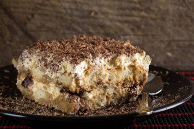 Tiramisu keki ve kaşık siyah tabakta, ahşap arka planda.