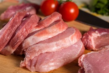 Ahşap kesim tahtasının üzerinde çiğ domuz eti, maydanoz ve arka planda domates dilimleri.