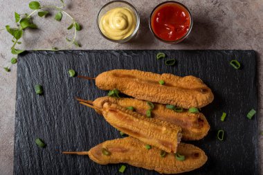 Domates sosu ve hardallı koyu bir tahtada dört taze mısır sosisi.