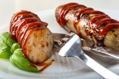 Sosisler ızgarada kızarır, ketçapla birlikte çatal ve bıçakla beyaz bir tabakta - yakın görüş