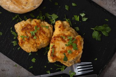 Koyu bir levhada iki parça Cordon Bleu ve bir parça ekmek ve doğranmış yeşil maydanoz.