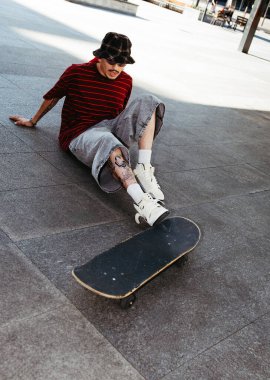 Genç bir kaykaycı şehir meydanında kayarken düştükten sonra yere oturur..