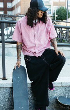 Genç patenci, kaykayını elinde tutarak, sokak kıyafetlerini giymiş bir şekilde raylarda oturuyor.