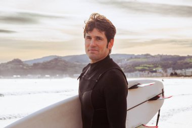 Dalgıç kıyafetli bir adam sörf tahtası tutuyor ve sahilde gülümsüyor.