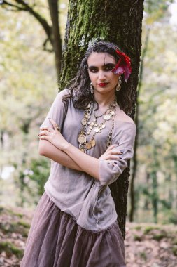 Zarif genç kadın ağaçların arasında poz veriyor, altın takılar ve tarafsız tonlar takıyor.