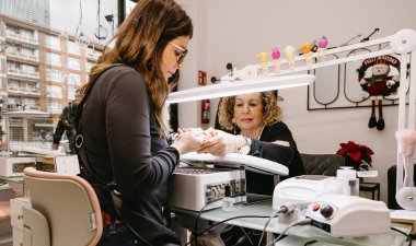 Bir manikürcünün, modern, parlak bir manikürcünün içinde olgun bir kadına manikür yapmasını geniş açıdan görmek. Görüntü arka planda temiz bir çalışma alanı ve şenlikli bir dekor gösteriyor.