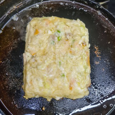 Altın kahverengili okonomiyaki, zengin lezzetli dokusunu, lahana karışımını ve geleneksel Japon sokak yemekleri tarzını gösteren bir elektrikli ocakta yemek pişirir..