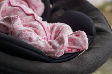 Detaylı bir yakın çekim, yumuşak, pembe, kelebek desenli bir tulum giyen bir bebeğin alt gövdesini yakalar. Bebek rahat bir şekilde yuvalanmış ve korunuyor. Karanlık, yastıklı bir taşıyıcı ya da bebek fedai.