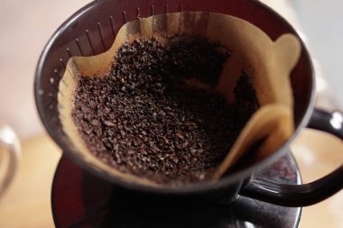 Detaylı bir çekim koyu, kaba kahve çekirdeklerini açık kahverengi kağıt filtrede dinlenirken gösteriyor. Siyah kahve damlatıcısının içine yerleştirilmiş, taze bir fincan demlemeye hazır..